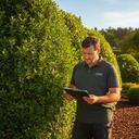 Landscape manager reviewing notes beside neatly trimmed hedges on a sunny job site